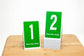 Tent style restaurant table numbers feature a bold white number on a green background. Please place visibly is printed at the bottom of each number.